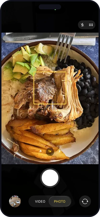 Phone camera framing a carnitas bowl with rice, black beans, plantains, and avocado
